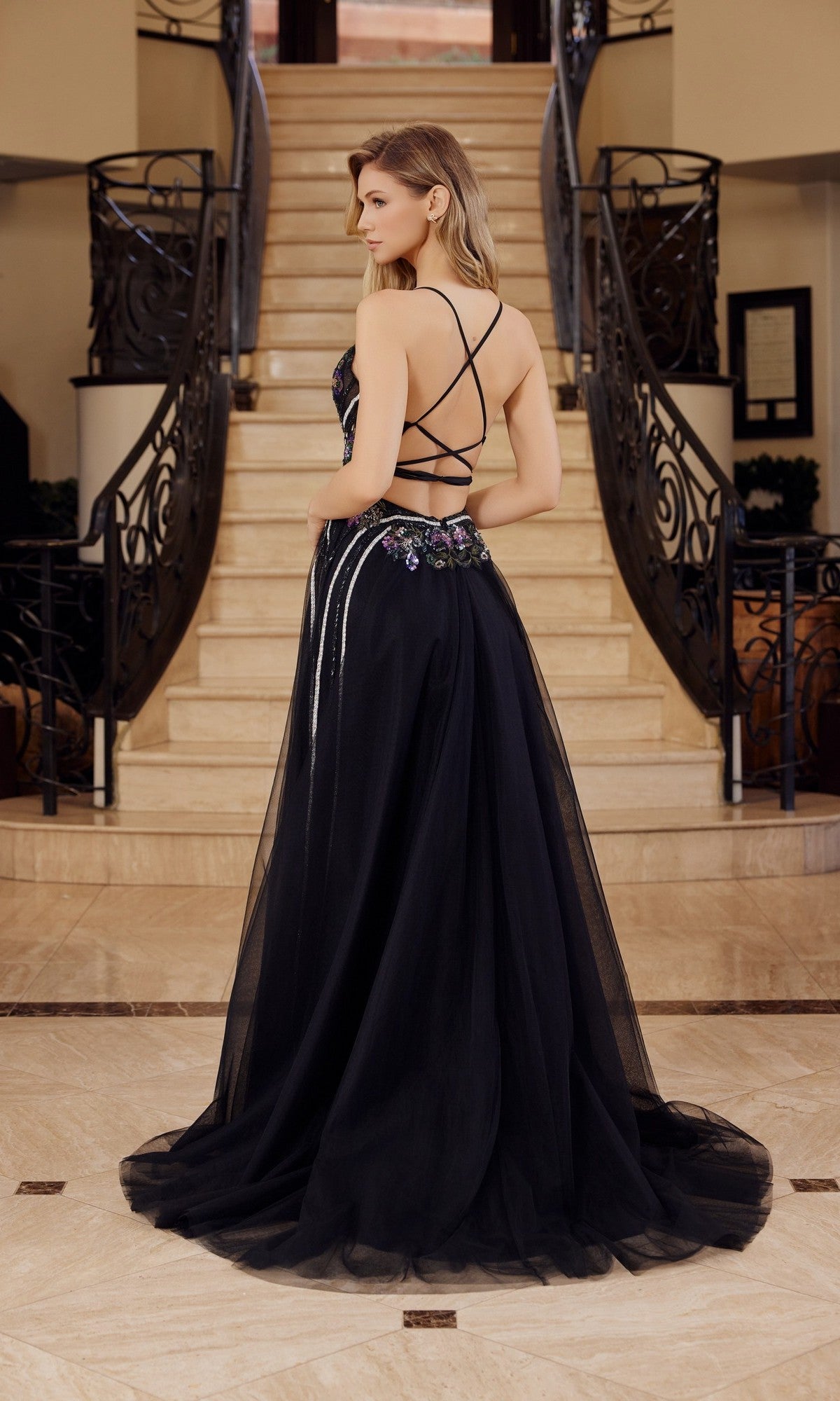 A woman wearing the Nox Anabel G1795 long prom dress, featuring a black open-back design with crisscross straps, stands at the base of a staircase facing away from the camera.